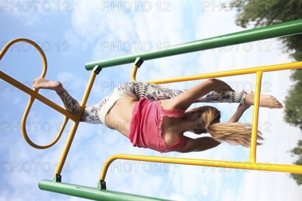 Athletic young blond gymnast working out on colourful metal bars in an outdoor playground hanging gracefully from one leg while stretching down with her other foot against a cloudy blue sky