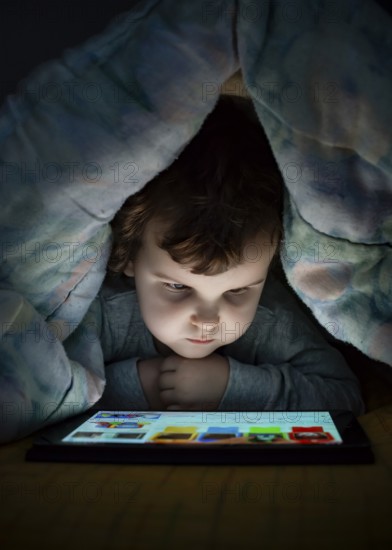 Little girl watching her tablet in the bed. Illuminated child face from device screen. Child dressed with pajamas under the covers hold a tablet. Night time
