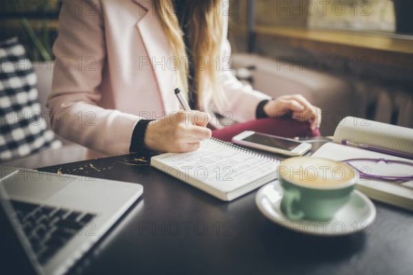 Close up caucasian woman hand on wooden table inside cafe makes notes in notebook. Subject freelancer blogger journalist at work. Unrecognisable person. Technology phone and laptop with cup coffee