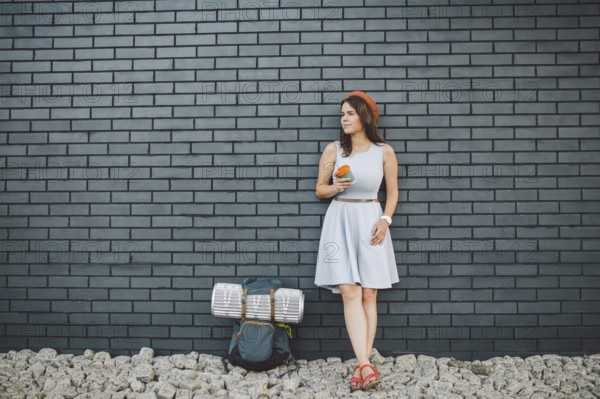 Cheerful girl in dress holding take away cup. Hipster model wearing stylish summer outfit Outdoors. woman with backpack walks along city street. hipster girl on brick wall, lifestyle of modern teenage
