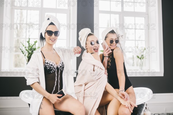 Theme female friendship. Party girlfriends, relaxation and spa procedures, hen party. Three Caucasian beautiful women sitting inside on bathroom in sunglasses, head towel and bathrobe, underwear