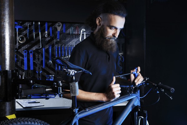 Theme sale and repair of bicycles. Young and stylish with a beard and long hair, a Caucasian man uses a tool to set up and repair a bike in a store. Business owner at work