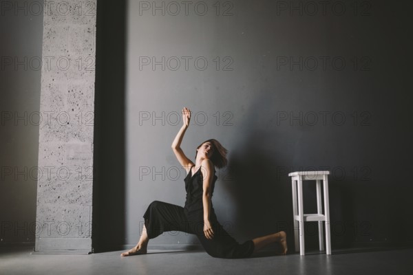 Social theme of female loneliness is pain suffering. Abastration of male domestic violence. Young beautiful Caucasian woman in black clothes, strange pose, white chair big dark room. Fear and despair