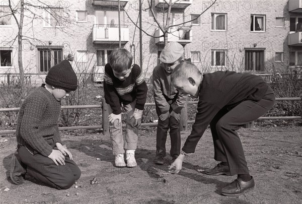 In the 1960s. Boys outdoors playing marbles. An old fashioned game where the goal is to hit the pyramid of marbles with just one marble. If you hit, you won the marbles in the pyramid, otherwise the marble was lost. There was also variants of the game. Sweden spring 1964. Roland Palm ref pärm 2