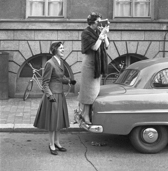 In the 1950s. An elderly woman with a film camera has stood on the bumper of a car to get a better view of what she is filming. A younger woman stands next to it, looking happy. They are relatives of one of the recently completed students who are celebrating that they have passed their matriculation exam and are walking down a Stockholm street. 1954. Svahn ref SVA2
