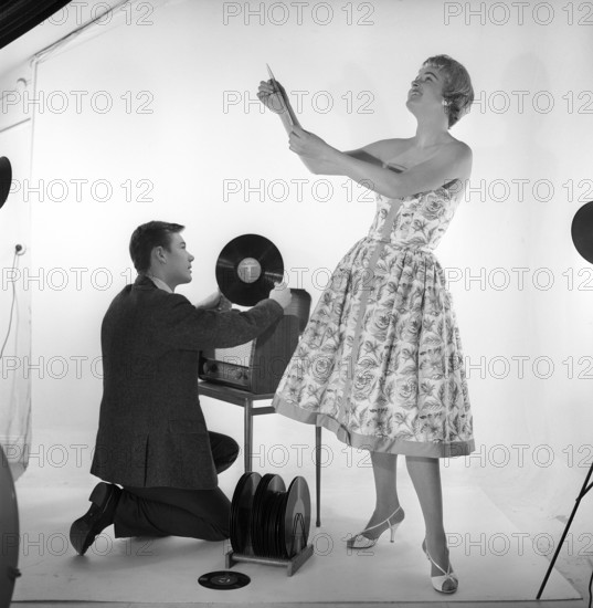 Gramophone in the 1950s. An man is kneeling by a combined recieving set and gramophone cabinet where he has opened the cover of the gramophone and holds a record in his hand.She is dressed in a typical 1950s dress looking happy. Sweden 1959. Kristoffersson ref CD65-2