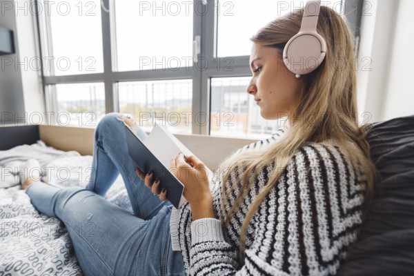 Blonde young caucasian woman lying on her dorm room bed next to a window putting on headphones, listening to music on a cold cloudy autumn day