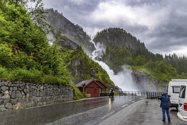 Woman tourist taking pictures of scenic double waterfall Lotefossen. Summer rainy day. The road passes through water jets. Cloud of water dust hangs in the air. Travel to Norway
