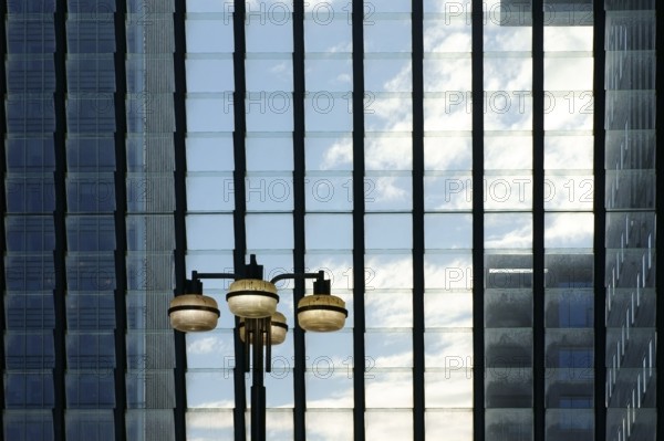 A glass façade with rows of hinged windows in front of which a lantern stands and tower blocks shine through in the background
