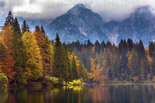 Lake Fuzine. Grand mountain range Dolomite Alps in thunderclouds. Border between Italy and Slovenia. Orange, yellow and red trees are reflected in the green smooth water of the lake