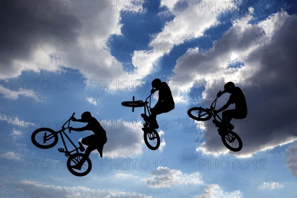 An extreme rider is making a free style jump from a ramp. The young boy with his bicycle is seen as a silhouette in front of the sun. Multiple exposure image. Clouds