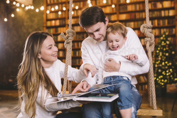 Family With Baby Relaxing On home Swing Seat in cozy dark living room on Christmas eve. Family having fun on seesaw. Winter evening with family and kid. Family Reading Book In Living Room