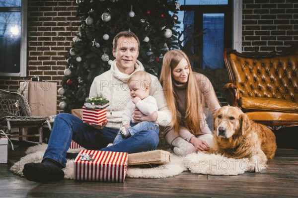 Theme Christmas and New Year family circle and domestic pet. Mom dad and child 1 year old Caucasian woman sitting on floor near Christmas tree with gifts and big dog breed labrador golden retriever