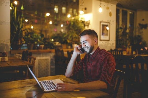 Young handsome Caucasian man with beard and toothy smile in red shirt works behind laptop, hands on keyboard sitting at wooden table. Uses calls on mobile phone. In evening at the coffee shop