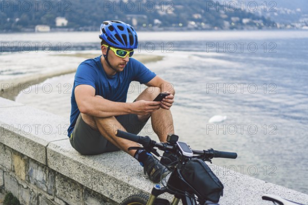 The theme of tourism and travel in Italy. A male cyclist uses a phone on the shore of Lake Como. Tourist guy in a helmet with a bicycle on the shore of a mountain lake in northern Italy