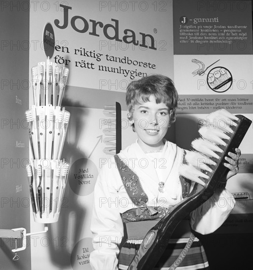 A young woman from the company Jordan is standing in their booth at an exhibition, holding a giant toothbrush. Behind her advertising the company's products and its functions. Alongside medium-hard and hard toothbrushes, as well as children's toothbrushes. Sweden 1952. Conard ref 4671