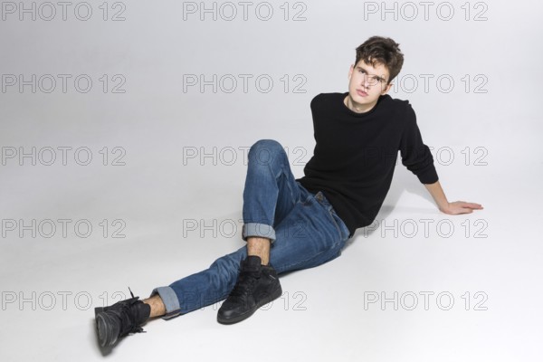 Stylish guy posing in studio sitting on the floor in denim pants and black sweatshirt. Male student portrait full on white background. Studio shot Handsome attractive european man sitting on floor