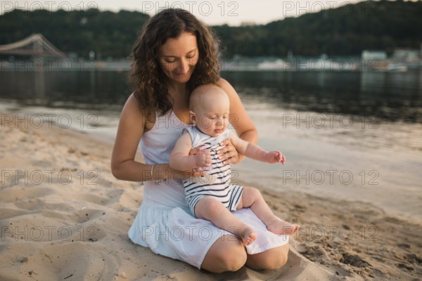 Young mother sitting on the beach with one year old baby son. Boy hugging, smiling, laughing, summer day. Happy childhood carefree game on the open sand, river lake, joy, fun. holiday concept