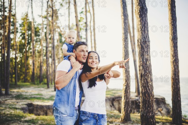 Happy young family of three people resting in a park outside the city. daughter sits at daddy on shoulders, and parents show direction forward by a hand for horizon.dressed in stylish denim clothes