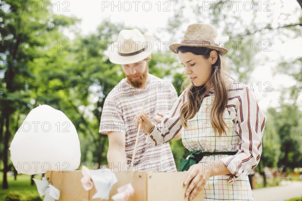 Theme is a family small business cooking sweets. A pair of lovers A young man and woman trader in the hat the owner of the outlet makes candy floß, fairy floß or Cotton candy in the park in summer