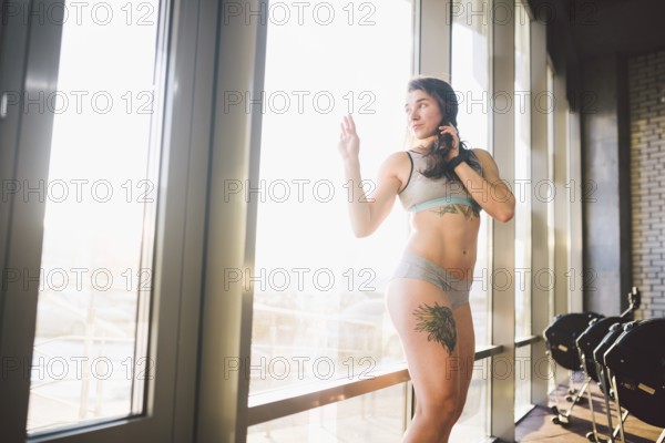 Young caucasian sporty woman with curly long hair poses in sportswear near a panorman window in a fitness club. Young female athlete model with beautiful body stands in the gym with a tattoo