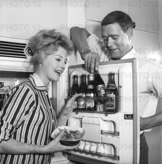 In the kitchen 1950s. A couple in their kitchen and at the refrigerator where the beer is kept in the door shelf. Five beer bottles are seen. Mrs is holding a bowl of shrimps. Sweden 1959. Kristoffersson ref CH77-1