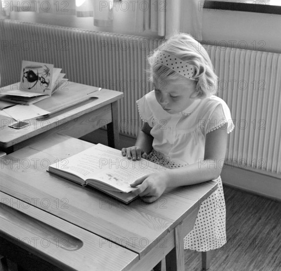 School in the 1950s. A girl is seen sitting at her desk in the classroom reading from the schoolbook in front of her. She is nicely dressed in a white skirt with a dotted pattern and matching headband. Sweden 1957 Conard ref 3538