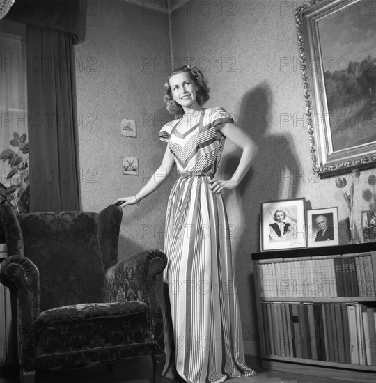 Fashion of 1946. A woman pictured wearing the typical fashion of the postwar era. She is wearing a long dress, the fabric of which is a pattern of narrow and wide fields of different colours. She is standing in a room by an armchair.Kristoffersson ref Y129-3
