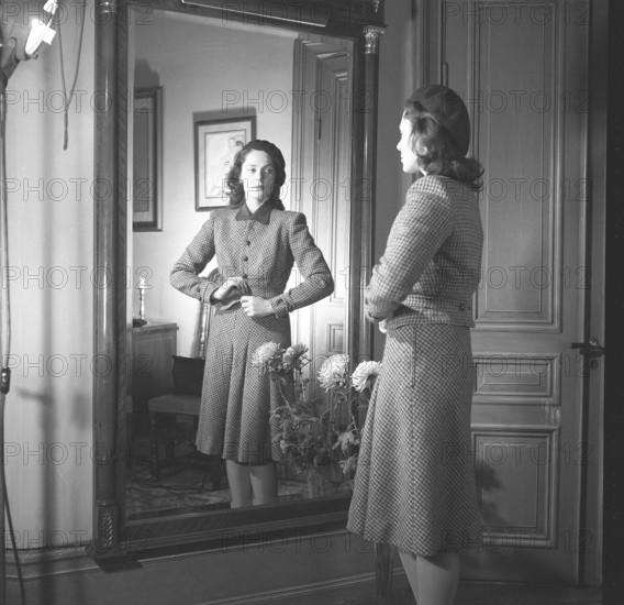 Actress Viveca Lindfors, 1920-1995. She stands in front of the mirror and buttons the last button of her plaid two-piece suit with a knee-length skirt and a waist-length jacket. 1945. Kristoffersson ref R55-6