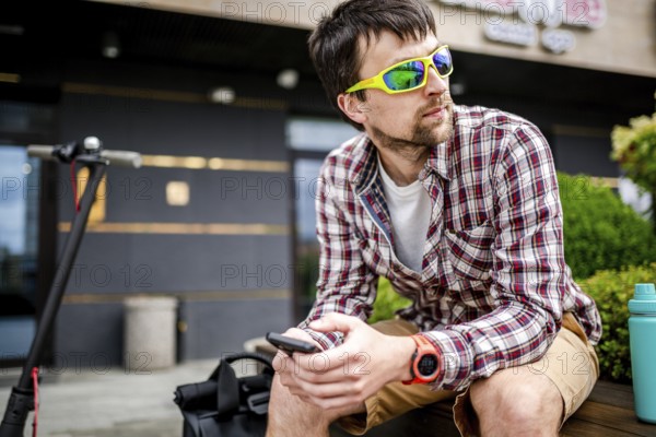 Individual urban environmental friendly transport electric kick scooter parked near man sitting on wooden bench in recreation area. Male use phone, surfing Internet, online shopping at smartphone