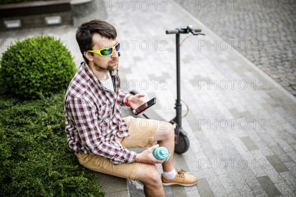Man with e-scooter relaxing and using smartphone on bench in city. Modern male with electro scooter sitting on bench in park while chatting on his mobile phone. Student with eco transport hold phone