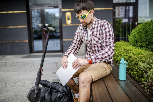 Young caucasian man in casual clothes freelancer uses eco-friendly electric transport scooter. Male takes out laptop from backpack, sitting on bench near building. Person unpacks laptop near office