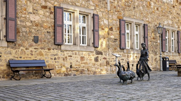 Scene in front of a stone building with shutters, a bench and a statue of a girl with geese, The Gänseliesel Monument in Melsungen