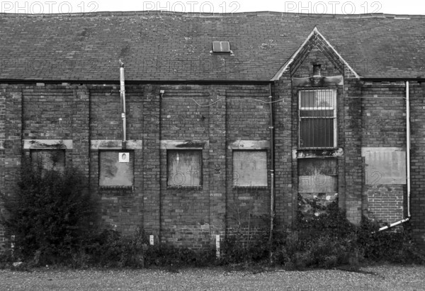 An old abandoned brick factory building with boarded up windows and crumbling decaying walls overgrown with weeds