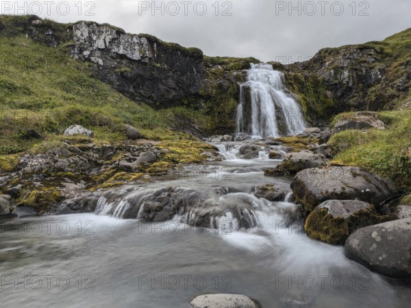 Waterfall cascading over mossy rocks with flowing water and greenery under a cloudy sky. Iceland