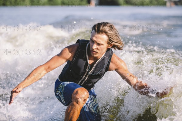 Beautiful long haired blond wakesurf in vest on board along waves of lake. Athletic male athlete wakes surfing in summer on river. Wakesurf water sport. Surfer on wave. Man surfing on the surfboard