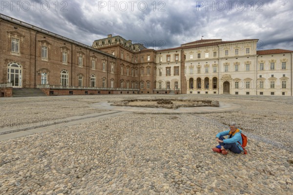 Woman sitting on a pebble square in front of a large building, Palace of Venaria Reale, Residences of the House of Savoy, Turin, Piedmont, Italy, Europe