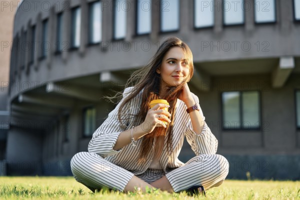 Young stylish woman drinking coffe and relax sitting on a grass in city park. Break time concept