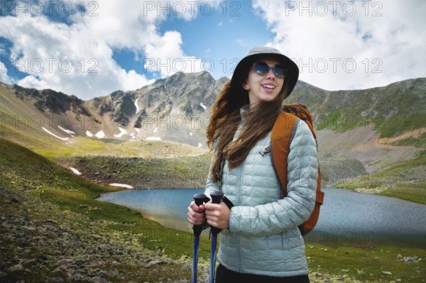 Smiling, positive girl with disheveled hair against the background of a blue mountain lake, with trekking poles in her hands and a backpack, a tourist on a hike