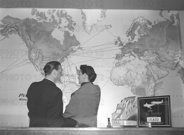In the 1950s. A young couple at a travel agency stands in front of a world map and looks at all the flight routes of the American airline Pan American between different countries and continents. Pan Am's classic logo is visible on the map and on the folder on the counter. The airline was the largest international airline in the US with the most international flights. 1955. Kristoffersson ref BX51-4