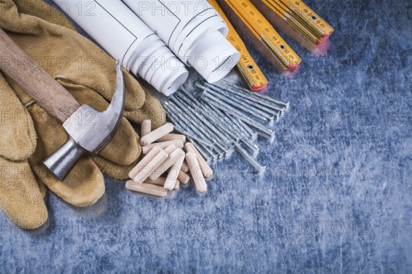 Blueprint rolls claw hammer safety gloves wooden meter metal nails and dowels on scratched metallic surface construction concept