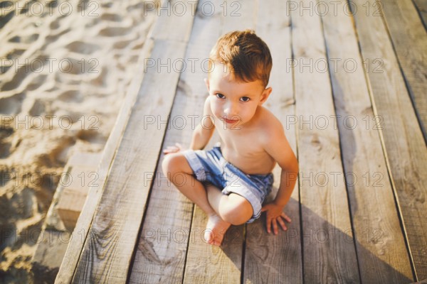 Little Caucasian boy child sitting on wooden pier sandy beach, summer time, sea holiday near water. The theme is the flow of time, a short life, the meaning and purpose of existence. Meaning of life