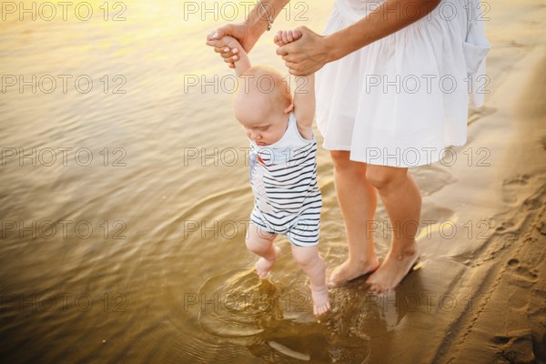 Baby boy walking along beach with mom's support. Mother teaching baby to walk. Outdoor, trust family concept. Mum Holding Baby For the First Step