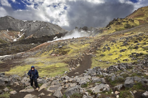 Rhyolite mountains smoldering underground heat. In the hollows lie unmelted snow patches from last year. Mature woman tourist wearing warm up the trail. Iceland in July