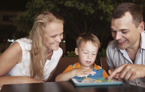 Young parents and little son in a cafe in the evening. Father and son using touch pad, mother watching them
