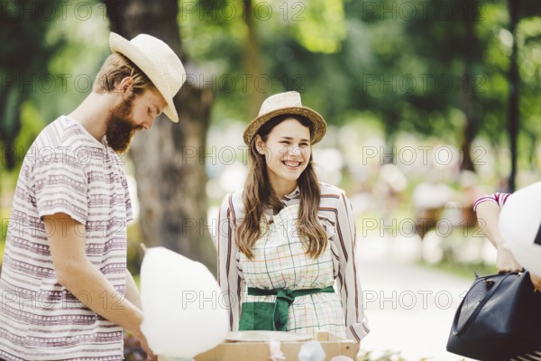 Theme is a family small business cooking sweets. A pair of lovers A young man and woman trader in the hat the owner of the outlet makes candy floß, fairy floß or Cotton candy in the park in summer