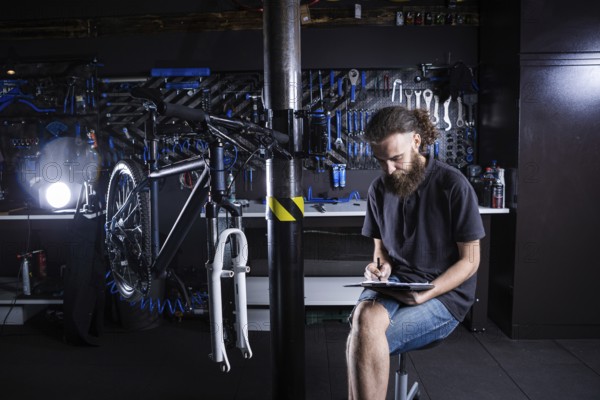 Stylish man with beard, cycle mechanic takes notes, checklist before repairing and servicing bike in bicycle shop workshop. Caucasian male in bicycle shop writing on clipboard. Assessing bike supply