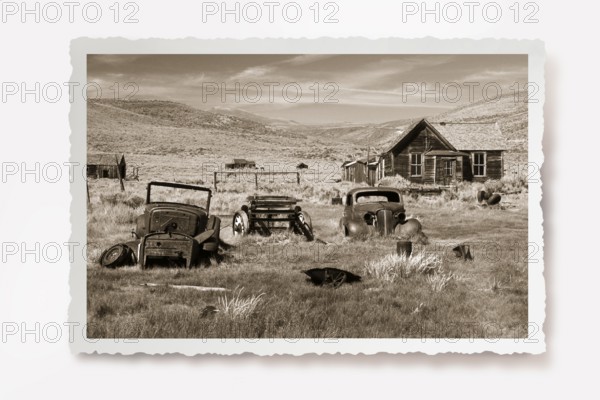 Several rusting car wrecks in a meadow in front of a wooden house in the former gold mining town of Bodie. Photo in sepia tone with white jagged edge, reclining shadow. Several rusting old car wrecks in a meadow in front of a wooden house in the former gold mining town of Bodie. Photo in sepia tone with white jagged edges