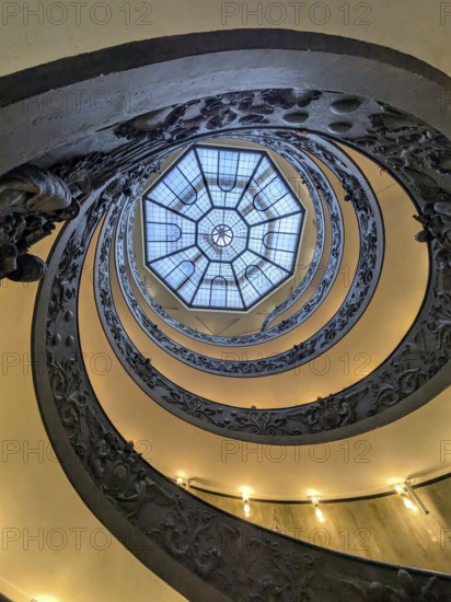 View from below of an ornate spiral staircase with a geometric skylight at the top, showcasing intricate metalwork. The Vatican