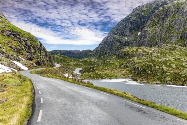 Narrow dangerous mountain road glitters from the rain. The roadside is marked with high poles. Traveling by camper on road 520. Gray rocks surround the lake. Summer in Norway